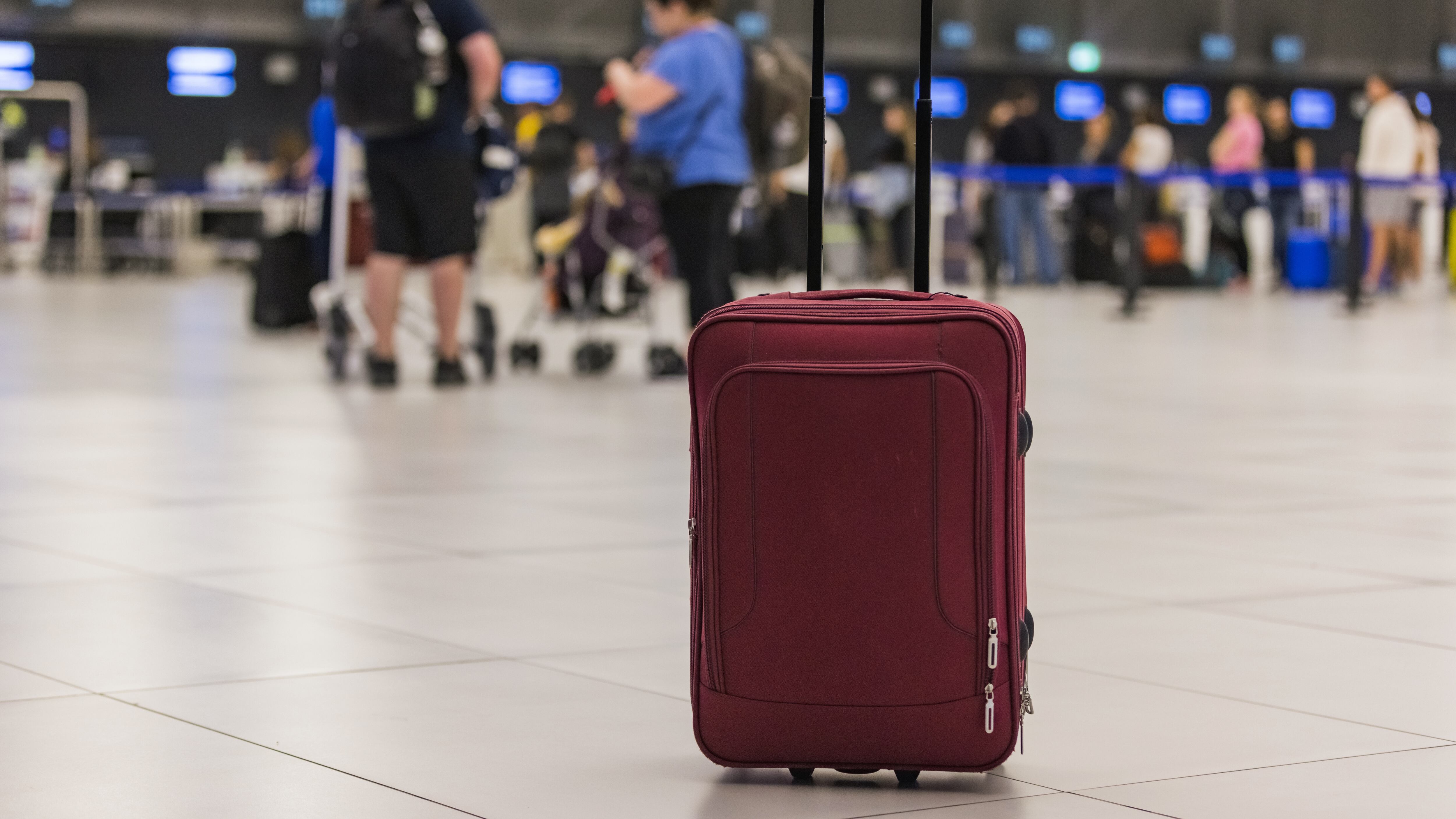 Red-brown rolling travel suitcase at the airport terminal with blurred passengers in the background.